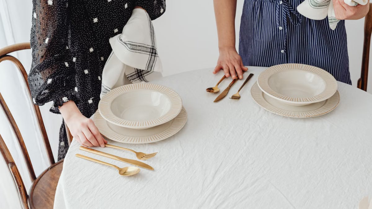 Two people setting a table with ceramic bowls and gold cutlery indoors.