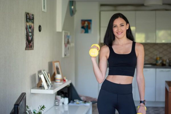 Smiling woman in activewear lifting dumbbells at home for a healthy lifestyle.
